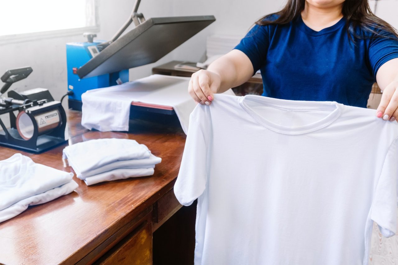 female-holding-a-white-t-shirt-next-to-the-cloths-printing-machine-and-t-shirts-on-the-wooden-table-1.jpg